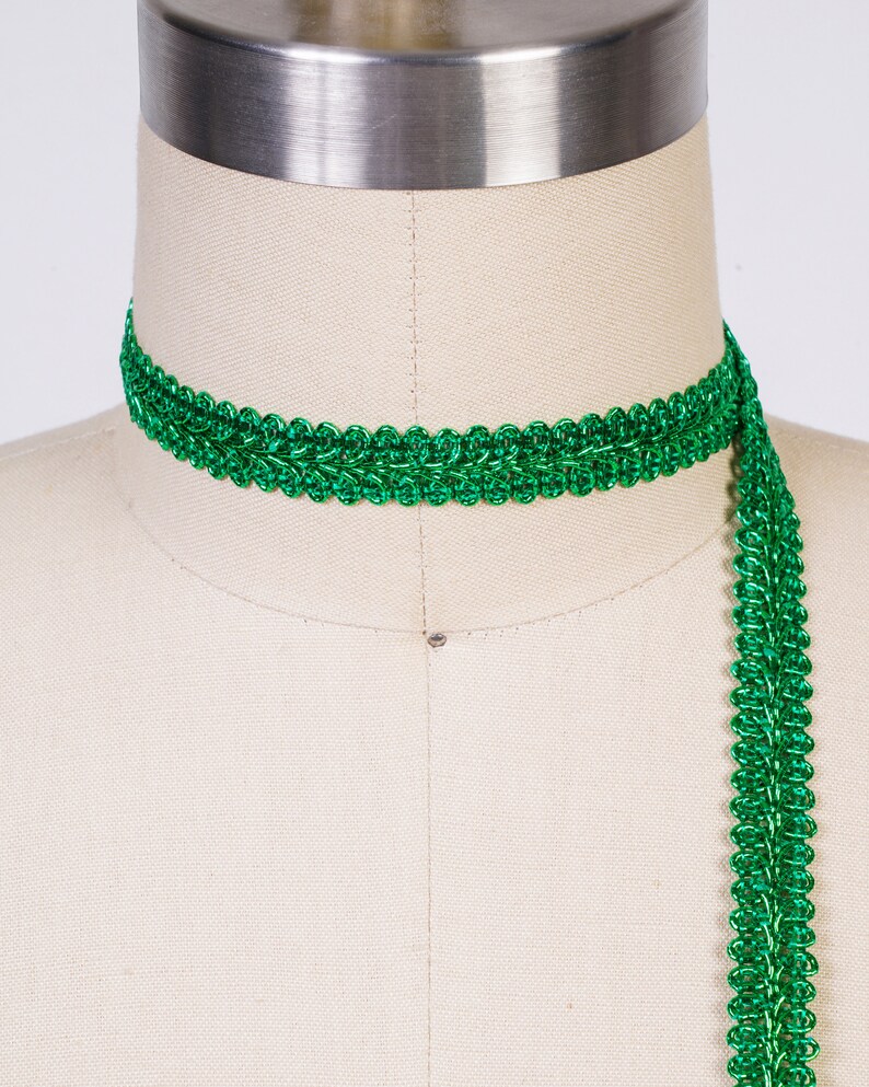 May include: Green metallic trim with a decorative woven pattern. The trim is shown wrapped around a dress form.