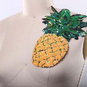 Può includere: Un'applicazione di paillettes a forma di ananas gialla e verde. L'ananas è fatta di paillettes e ha una parte superiore verde con foglie.