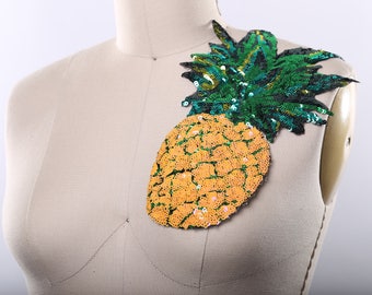 Może przedstawiać: Naszywka w kształcie ananasa z żółtych i zielonych cekinów. Ananas jest wykonany z cekinów i ma zieloną, liściastą górę.