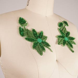 May include: Two green floral appliques with sequins, each featuring a large flower with smaller leaves and buds. The appliques are displayed on a white fabric background.