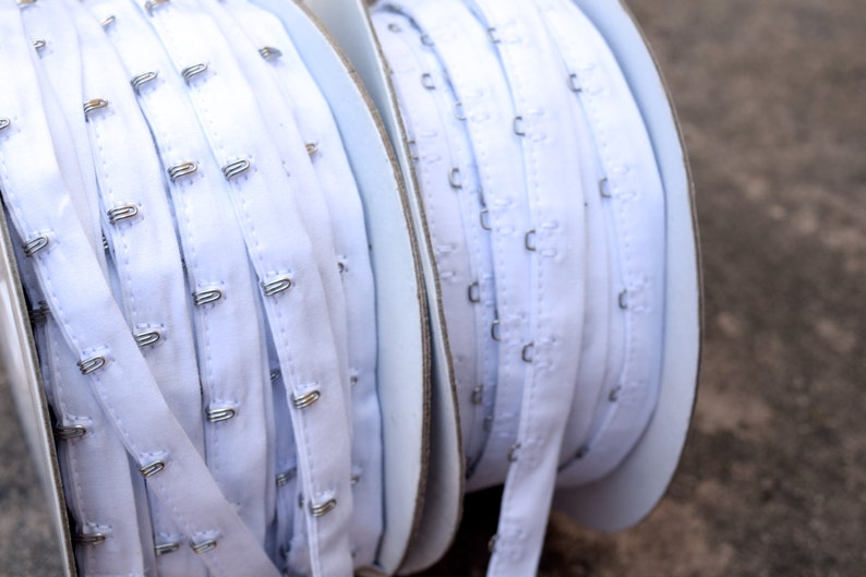 May include: Two spools of white elastic bra strap with metal hooks. The spools are on a white background.