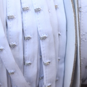 May include: White bra strap with silver hook and eye closures. The strap is folded and rolled up, showing multiple rows of hooks and eyes.