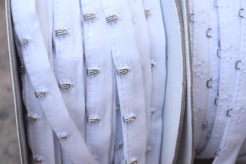 May include: White bra strap with silver hook and eye closures. The strap is folded and rolled up, showing multiple rows of hooks and eyes.