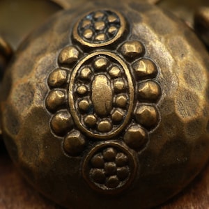 Pode incluir: Um close-up de um botão de metal dourado com textura martelada e detalhes decorativos. O botão é circular e apresenta três círculos concêntricos de pontos em relevo.