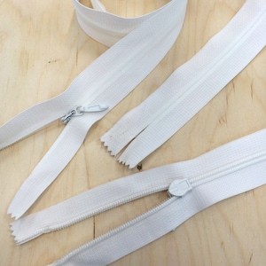 May include: Three white zipper closures, two with open ends and one with a closed end. The zippers are laid out on a light wood surface.