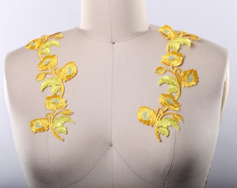 May include: Two yellow floral appliques with green leaves and silver glitter accents. The appliques are on a white fabric background.