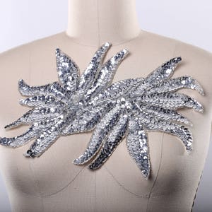 May include: A silver sequined applique in the shape of two stylized flowers. The applique is perfect for adding a touch of sparkle to clothing, bags, or home decor projects.