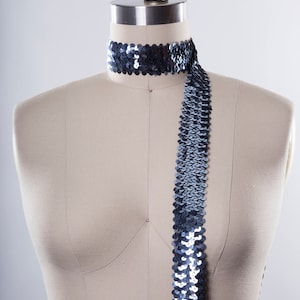 May include: A silver sequined trim with a slight iridescent sheen. The trim is draped over a dress form.