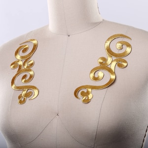 May include: Two gold embroidered fabric appliques in a floral design. The appliques are on a white fabric background.