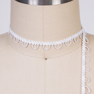 May include: White elastic trim with a scalloped edge. The trim is shown on a dress form.