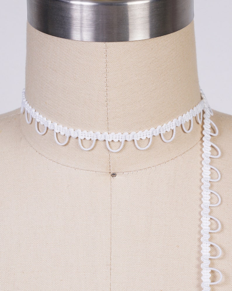 May include: White elastic trim with a scalloped edge. The trim is shown on a dress form.