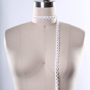 May include: A white fabric covered dress form with a white scalloped trim around the neck and a long piece of white scalloped trim hanging down the front.