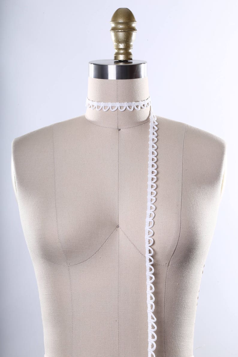 May include: A white fabric covered dress form with a white scalloped trim around the neck and a long piece of white scalloped trim hanging down the front.
