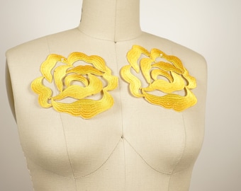 May include: Two large, yellow, embroidered floral appliques in the shape of roses. The appliques are on a white fabric background.