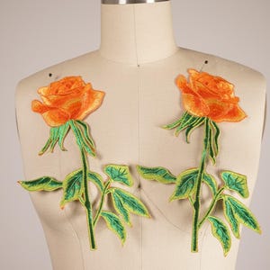May include: Two orange embroidered rose patches with green leaves and stems. The roses are in full bloom and have a delicate, intricate design.