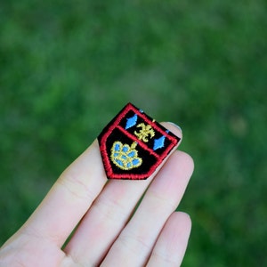 May include: A black embroidered patch with a red border. The patch features a gold and blue design with a crown and a stylized flower.