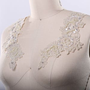 May include: A pair of white lace appliques with pearl embellishments. The appliques are designed to be sewn onto a garment, such as a wedding dress or bridal gown.