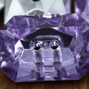 May include: Close-up of a faceted, lavender-colored button with two holes. The button has a geometric design and a shiny, reflective surface. Other buttons are visible in the background.