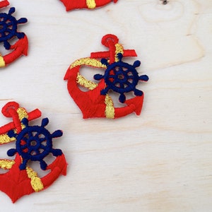 Puede incluir: Cuatro parches de ancla bordados en rojo y dorado con timones azules sobre un fondo de madera clara.