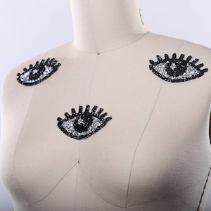 May include: A white fabric mannequin with three black and silver eye appliques. The eyes are sewn onto the fabric of the mannequin's chest and shoulders.