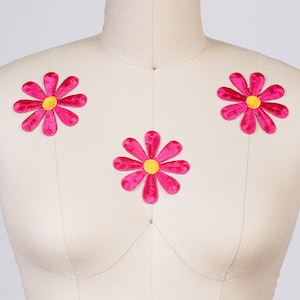 May include: Three pink felt flower appliques with yellow centers. The flowers are attached to a white fabric background.