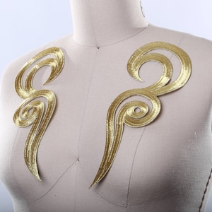 May include: Two gold embroidered fabric appliques in a swirling, abstract design. The appliques are on a light beige fabric background.