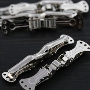 Puede incluir: Dos pinzas para el pelo de metal plateado, una abierta y otra cerrada, sobre una superficie de madera oscura. Las pinzas están diseñadas para sujetar el pelo en su lugar.