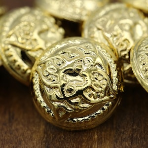 May include: A close-up of a pile of gold-colored metal buttons with intricate designs. The buttons are round and have a raised, textured surface.