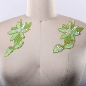 May include: Two green floral appliques with sequins, each featuring a five-petal flower with a pearl center. The appliques are attached to a light brown fabric background.