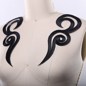 Peut inclure: Deux appliques en tissu noir dans un design abstrait tourbillonnant. Les appliques sont sur un fond de tissu blanc.