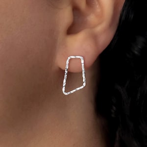 Peut inclure: Une boucle d'oreille clou en argent présentant un cadre géométrique ouvert et irrégulier avec une surface martelée texturée. La boucle d'oreille a une forme trapézoïdale aux coins arrondis et une finition métallique polie qui reflète la lumière.
