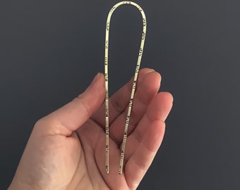 May include: A U-shaped gold-toned metal hair pin featuring a series of small, evenly spaced rectangular cutouts along both prongs and the curved top. The accessory has a smooth, polished finish and a slender, minimalist design for securing hair.