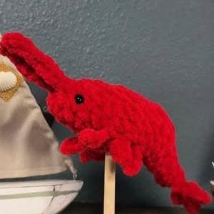 May include: A vibrant red, crocheted shrimp toy with a long snout and small black eyes. The shrimp is mounted on a wooden stick, positioned in front of a blurred background with miniature sailboat decorations.
