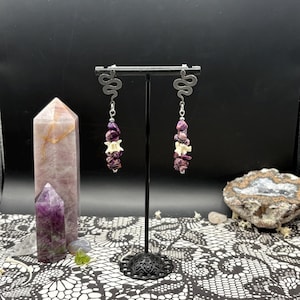May include: Dangle earrings with a snake design at the top, featuring a combination of purple and white stones. The earrings are displayed on a black stand against a black backdrop. The earrings are a unique accessory.
