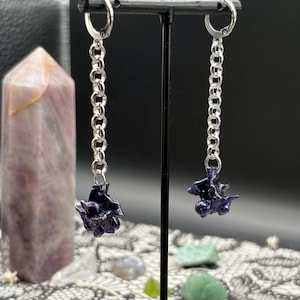 May include: Silver chain earrings with dark purple, textured charms. The earrings hang from silver hoops and are displayed on a black stand. A purple crystal and small gemstones are in the background.