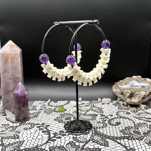 May include: A pair of hoop earrings with a unique design. Each earring features a white, bone-like structure and is accented with three purple, spherical beads. The earrings are displayed on a black stand against a dark background.