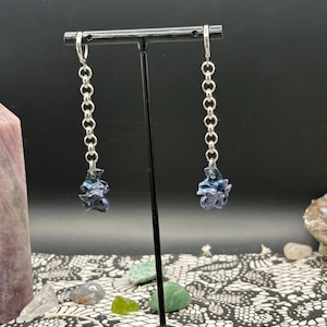 May include: A pair of dangle earrings with silver-colored chain links and abstract, dark blue and purple resin pendants. The earrings are displayed on a black stand against a dark background.