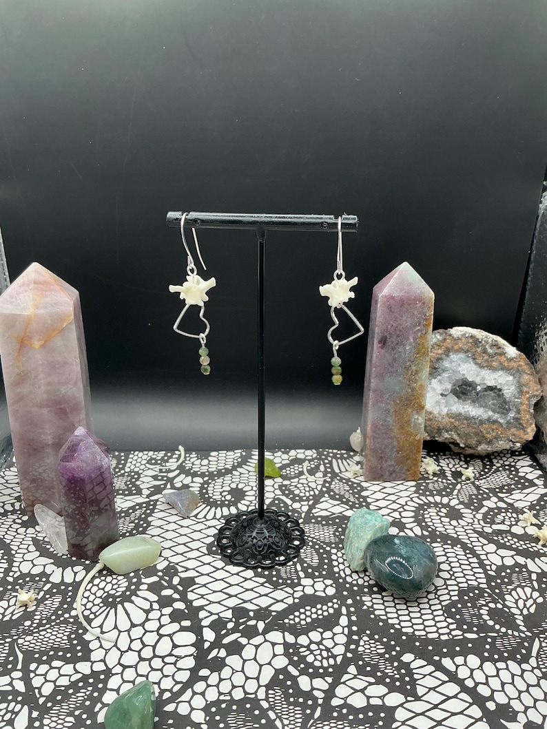 May include: A pair of silver-toned earrings featuring small white bone vertebrae segments, silver wire heart-shaped frames, and dangling green gemstone beads, suspended from a black metal T-shaped display stand.