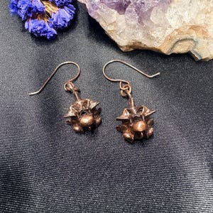 May include: A pair of copper-colored earrings with a unique, textured design. The earrings have a floral or starburst-like shape, with intricate details. The hooks are simple and curved. The earrings are displayed on a dark, textured surface.