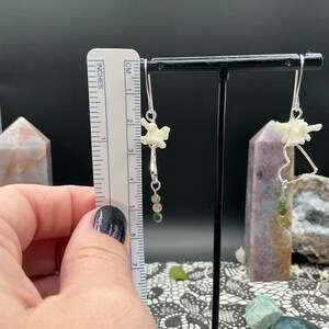 May include: A pair of dangle earrings featuring small white bone vertebrae, silver-toned heart-shaped wire frames, and green beaded accents. A ruler shows the earrings measure approximately 2.25 inches in total length from the top hook to the base.