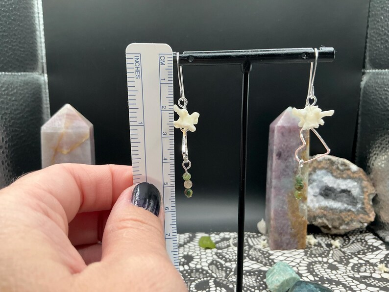 May include: A pair of dangle earrings featuring small white bone vertebrae, silver-toned heart-shaped wire frames, and green beaded accents. A ruler shows the earrings measure approximately 2.25 inches in total length from the top hook to the base.