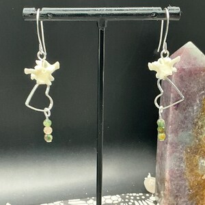 May include: A pair of dangle earrings featuring silver-toned hooks, a small white bone vertebra segment, a silver-toned heart-shaped wire frame, and a vertical strand of three small, round, multi-colored green and brown beads at the bottom.
