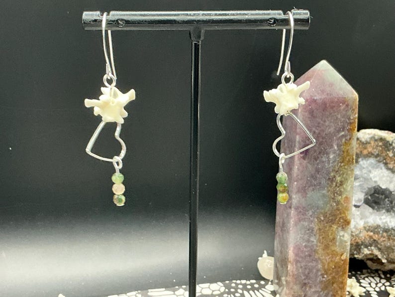 May include: A pair of dangle earrings featuring silver-toned hooks, a small white bone vertebra segment, a silver-toned heart-shaped wire frame, and a vertical strand of three small, round, multi-colored green and brown beads at the bottom.