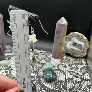 May include: A silver-toned earring featuring a small white bone-like segment and a dangling green bead, positioned next to a white ruler showing a length of approximately 1.5 inches from the top hook to the bottom of the green bead.