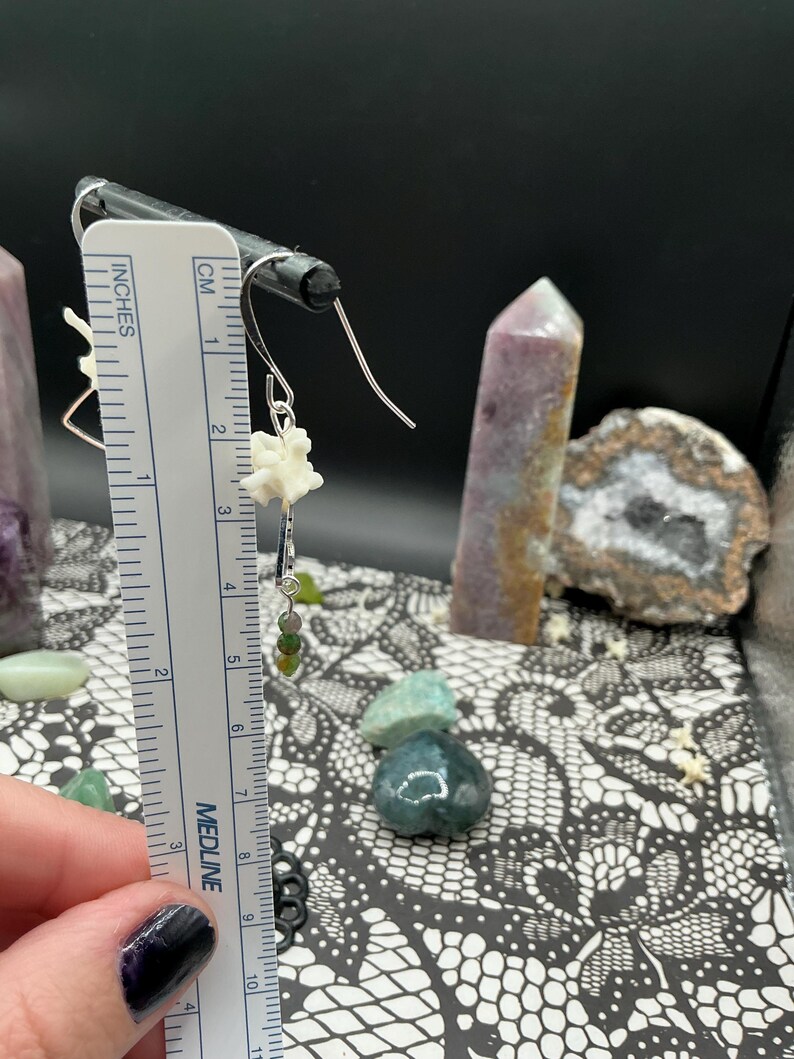 May include: A silver-toned earring featuring a small white bone-like segment and a dangling green bead, positioned next to a white ruler showing a length of approximately 1.5 inches from the top hook to the bottom of the green bead.