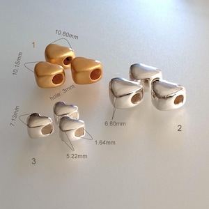 May include: A collection of heart-shaped beads in gold and silver tones. Group 1 features matte gold beads measuring 10.80mm by 10.15mm with 3mm holes. Group 2 shows shiny silver beads at 6.80mm. Group 3 displays smaller silver beads at 7.13mm by 5.22mm.