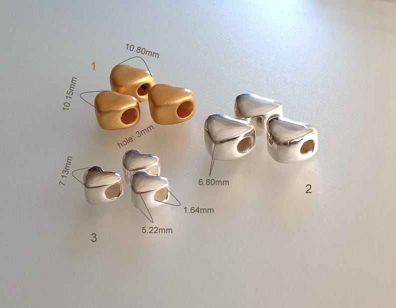 May include: A collection of heart-shaped beads in gold and silver tones. Group 1 features matte gold beads measuring 10.80mm by 10.15mm with 3mm holes. Group 2 shows shiny silver beads at 6.80mm. Group 3 displays smaller silver beads at 7.13mm by 5.22mm.