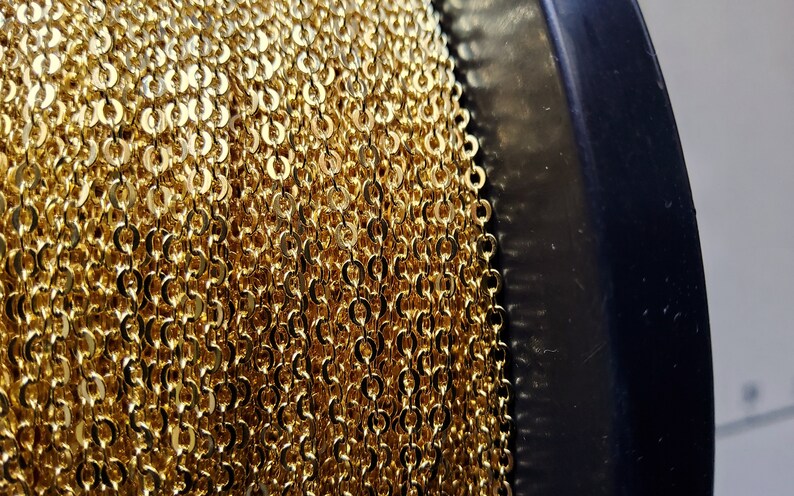K&ouml;nnte beinhalten: Nahaufnahme einer Spule mit goldfarbener Kette. Die Kette besteht aus kleinen, kreisf&ouml;rmigen Gliedern, die eng aneinander gewickelt sind. Die Kette hat eine helle, gl&auml;nzende Goldfarbe, und die Spule ist schwarz.