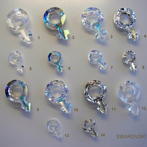 May include: Fourteen crystal charms in the shape of a female symbol, with iridescent rainbow colors. The charms are arranged in rows on a white surface. The word "SWAROVSKI" is printed in black at the bottom right corner of the image.