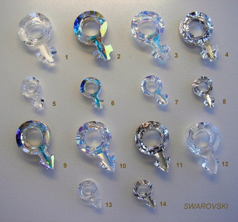 May include: Fourteen crystal charms in the shape of a female symbol, with iridescent rainbow colors. The charms are arranged in rows on a white surface. The word "SWAROVSKI" is printed in black at the bottom right corner of the image.